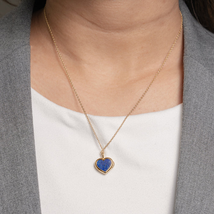A gold heart-shaped pendant necklace with a blue lapis lazuli stone, worn by a model in a gray blazer over a white top.