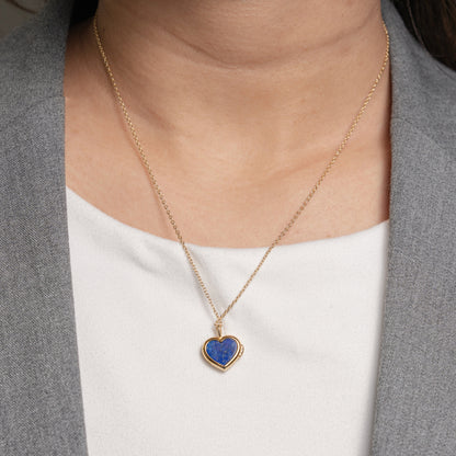 A gold heart-shaped pendant necklace with a blue lapis lazuli stone, worn by a model in a gray blazer over a white top.